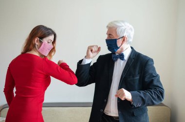 Olgun, gri saçlı bir adam ve kırmızı saçlı, maskeli bir kadın dirseklerini selamlıyor. El sıkışmaya ve sarılmaya bir alternatif. Meslektaşlar aralarındaki mesafeyi koruyorlar. Popüler bir selamlama şekli..