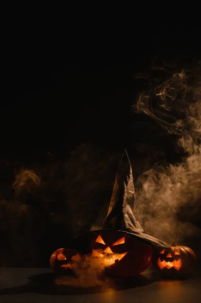 Tarjeta vertical de Halloween. Sombrero de bruja en una calabaza con muecas espeluznantes talladas sobre un fondo negro en la niebla. Jack-o-linterna brilla desde el interior y emite humo. — Foto de Stock