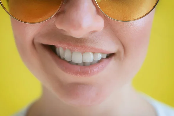 Närbild av en kvinnlig leende på en gul bakgrund. Beskuren bild. Kvinna med ett perfekt leende i solglasögon. — Stockfoto
