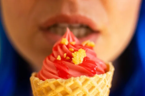 Closeup of sensual female lips with a waffle cone of pink ice cream. A cropped photo of a woman with blue hair enjoys eating pink gelato on a hot summer day. Macro. Happy sweet tooth.