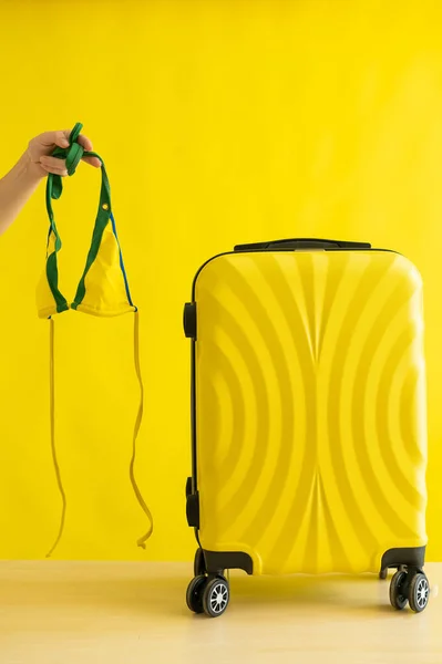 얼굴없는 여자가 노란 배경 여행 가방 옆에 비키니를 들고 여행을 갑니다. 한 여성 손이 여름휴가를 위해 수영복을 준비하고 있다. 복사 공간. — 스톡 사진