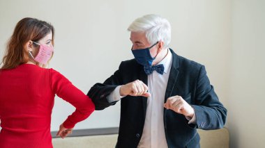 Takım elbiseli olgun, gri saçlı bir adam ve kırmızı elbiseli, tıbbi maske takan kırmızı saçlı bir kadın sosyal mesafeyi koruyup dirseklerini selamlıyorlar. El sıkışmaya ve sarılmaya bir alternatif..