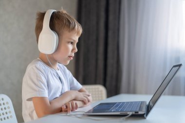 Sevimli çalışkan çocuk klavyede yazı yazıyor ve dizüstü bilgisayara dikkatlice bakıyor. Beyaz bir öğrenci bir masaya oturur, kulaklıkla müzik dinler ve ödev yapar. Uzaklık eğitimi.