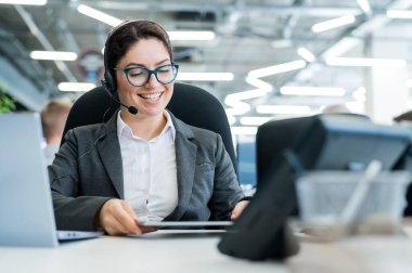 Mutlu bir bayan destek operatörü bir masada oturuyor ve telefonlara cevap veriyor. Güzel, gülümseyen bir kadın kulaklıkla müşterilerle konuşuyor. Kulaklıklı bir ofis çalışanı..