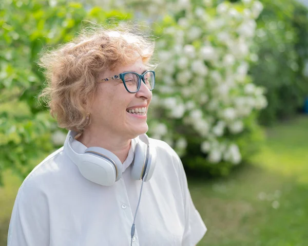 Une femme caucasienne âgée marche dans un parc et écoute de la musique. Une pensionnée souriante enlève son casque et profite de l'arôme des arbres à fleurs dans le jardin. — Photo