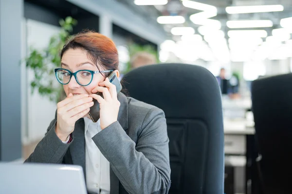 Una mujer se sienta en una oficina en su escritorio y chismorrea por teléfono. Ética corporativa. La empleada de traje cuenta secretos en un smartphone en el trabajo . —  Fotos de Stock