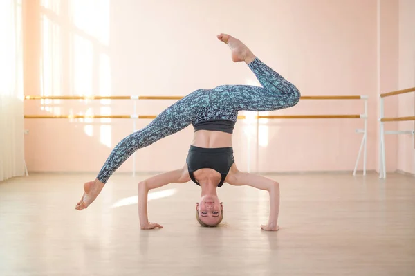 Flexibel vit flicka står på huvudet i en dansklass. Flickan gör yoga. . Kvinna dans breakdance på bakgrunden av balett barre. — Stockfoto