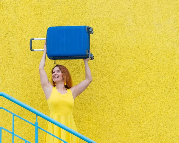 Happy woman in a yellow dress holds a blue suitcase over her head against the background of a yellow wall. Caucasian girl smiles with teeth and happily waits for a trip.