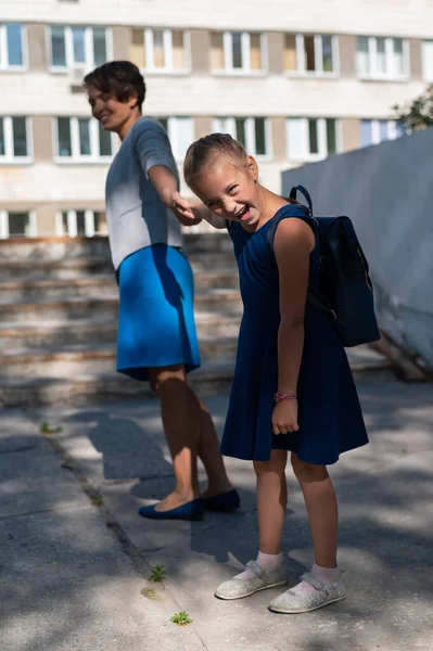 Маленькая несчастная девочка с рюкзаком тянет свою мать за руку и не хочет идти в школу. Женщина ведет сопротивляющуюся дочь в школу. Конфликт поколений. — стоковое фото