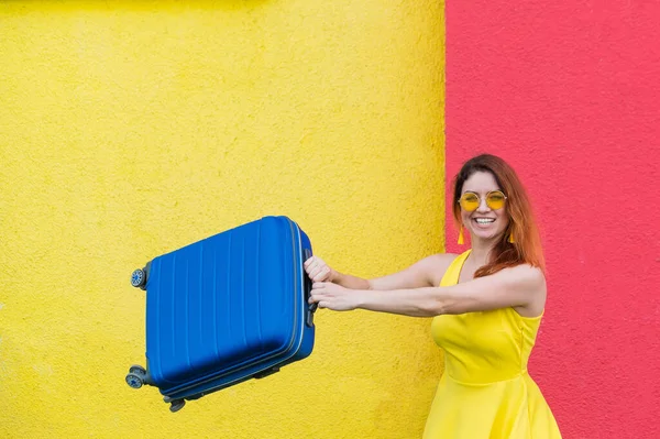 Šťastná žena v biči na žlutočerveném pozadí radostně zamává kufříkem v očekávání cesty. Krásná dívka ve slunečních brýlích s modrým sáčkem v rukou. Zavazadla pro letní dovolenou. — Stock fotografie