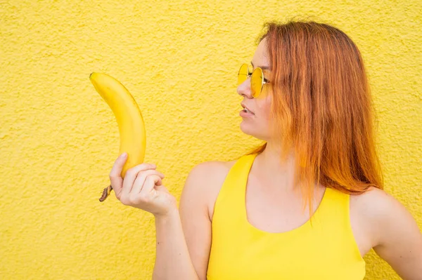 Porträtt av den kaukasiska rödhåriga kvinnan i solglasögon med en banan i händerna. Vacker förförisk flicka poserar på en gul bakgrund. — Stockfoto