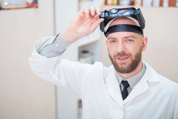 Maschio medico barbuto in un cappotto medico e con una cinghia di ingrandimento w luci seduto sulla scrivania. Attrezzature optometriche. Ufficio medico. — Foto Stock