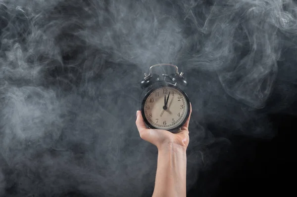 Close-up of a female hand holding a clock on a black background in smoke. Alarm clock at midnight in a mystical fog.