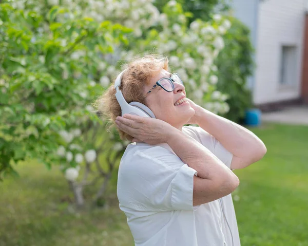 Una anciana caucásica camina en un parque y escucha música. Una abuela sonriente disfruta del aroma de los árboles florecientes en el jardín en un cálido día soleado . — Foto de Stock