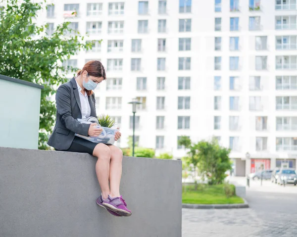 Depressed woman in medical mask sits on parapet outdoors with a box of personal items from the desktop. Unemployed girl alone on the street. Bankruptcy of a company due to quarantine in coronavirus.