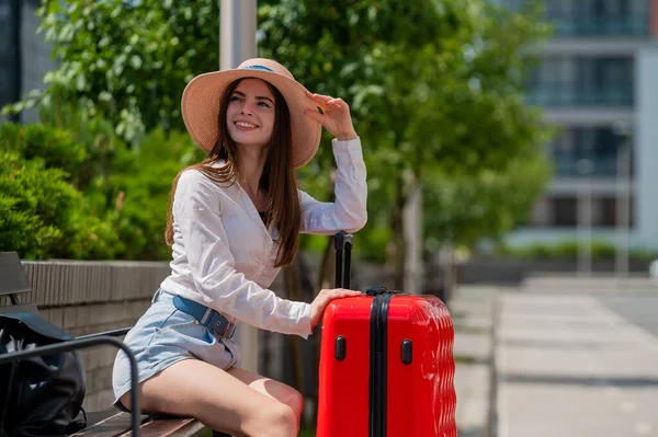 Eine schöne kaukasische Frau mit Hut und kurzen Hosen sitzt auf einer Bank und hält einen roten Koffer in der Hand und wartet auf ein Taxi oder einen Bus. Mädchen wartet auf Abreise auf Sommerreise. — Stockfoto