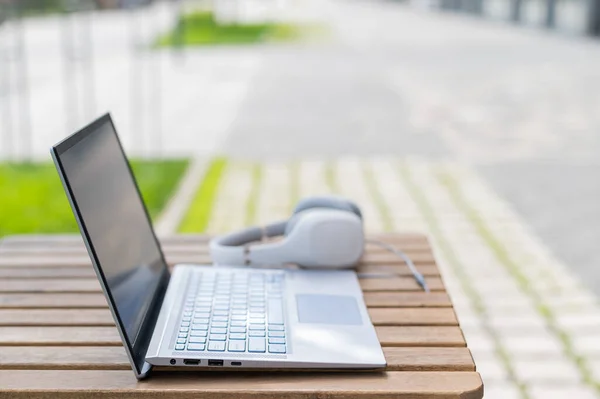 Close-up of a laptop and white big headphones on a wooden table on a summer terrace. No people. Cafe at the outdoors. Freelancer workspace. — Stock Photo, Image