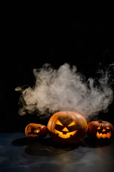 Drie griezelige Halloween stomende pompoenen met een gesneden lichtgevende grijns op een zwarte achtergrond. Een handgemaakte jack-o-lantaarn hoofd met een kaars in het donker tussen de mist. Snoep of ik schiet. Briefkaart. — Stockfoto