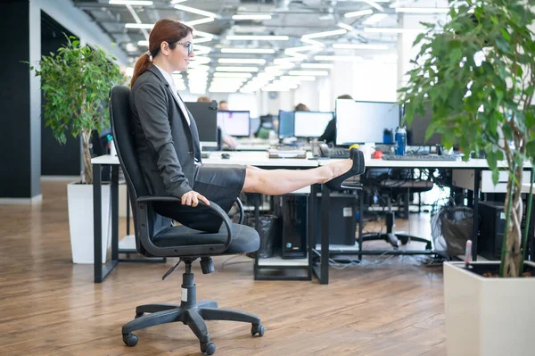 Happy business woman doing abdominal muscle exercises in an open space office. A red-haired smiling female employee in a skirt and high heels is dying off a chair in the workplace. Fitness at work.