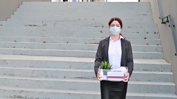 Ongelukkige vrouw met een masker loopt langs de straat met een doos persoonlijke spullen op de achtergrond van de trap. Een vrouwelijke kantoormedewerker werd ontslagen. Economische crisis tijdens epidemische hevige 19. — Stockvideo