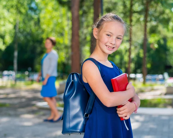Das Porträt einer fleißigen Schülerin mit Rucksack und Buch geht in die erste Klasse. Mutter schickt glückliche Tochter in die Grundschule. — Stockfoto