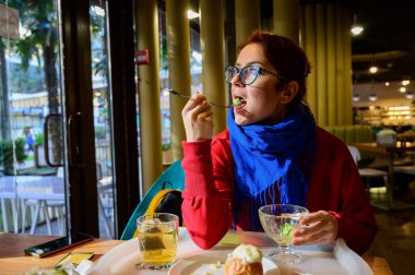 Kafede salata yiyen kızıl saçlı beyaz bir kadın. Gözlüklü ve mavi eşarplı düşünceli romantik kız yemek yiyor..