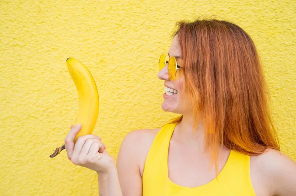 Portrét bělošské zrzavé ženy ve slunečních brýlích s banánem v rukou. Krásná svůdná dívka pózující na žlutém pozadí. — Stock fotografie