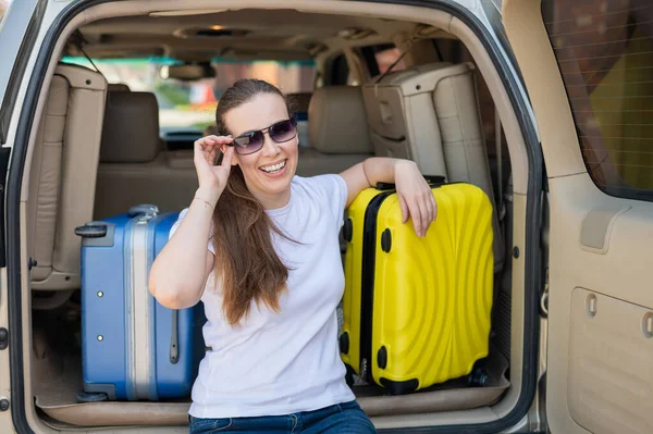 Mujer europea en gafas de sol empaca sus maletas para un viaje en coche. Hermosa chica feliz sentado en el maletero de un coche y listo para viajar. Maleta amarilla con cosas para las vacaciones de verano . — Foto de Stock