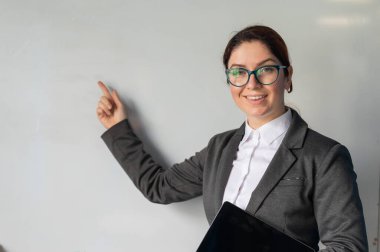 Elinde dijital tabletle takım elbiseli güzel kızıl saçlı kadın. Gözlüklü gülümseyen bir bayan öğretmen bir okul sınıfında duruyor ve beyaz bir tahtayı işaret ediyor. Eğitim Kurumu.