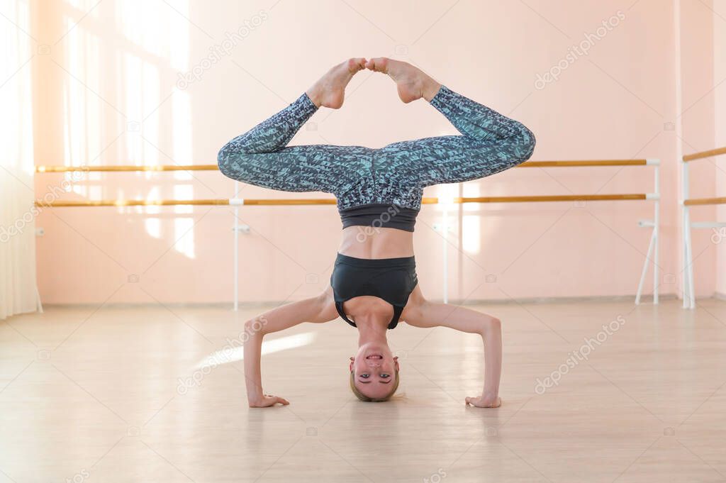 Flexible Caucasian girl stands on her head in a dance class. The girl does yoga. . Woman dancing breakdance on the background of ballet barre.