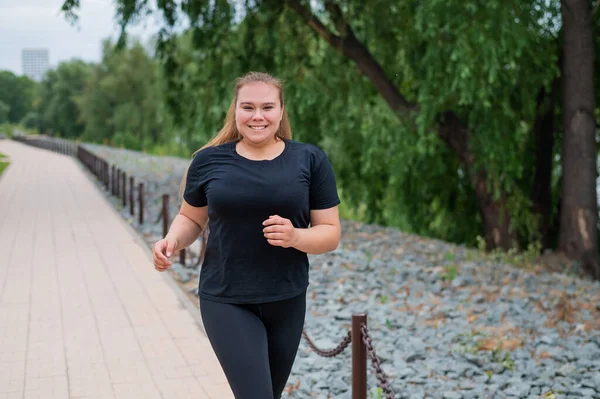 Жирна молода жінка біжить на вулиці. Товста красива усміхнена дівчина в чорному костюмі займається пристосуванням для схуднення на набережній. Жінка біжить в літній день . — стокове фото