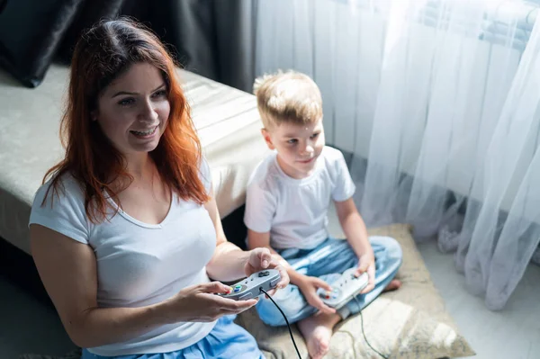 Happy woman and son play video games at home. A gambling boy and his mother are playing with the game console joysticks in their hands. A happy family.