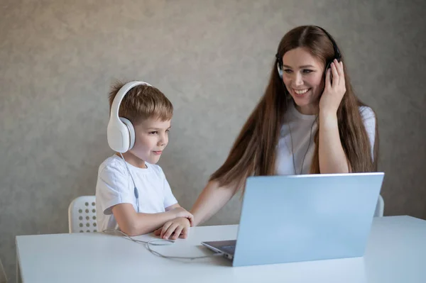 Szczęśliwa kobieta z synem w słuchawkach słuchająca muzyki na laptopie. Biały chłopiec z matką siedzą przy stole i patrzą na ekran komputera.. — Zdjęcie stockowe