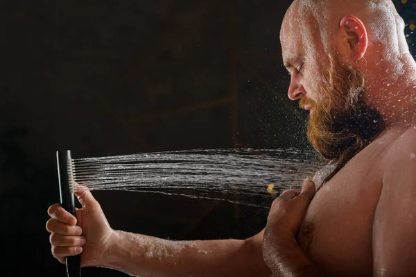 El macho toma una ducha. Un hombre brutal con barba roja está parado en el baño bajo el agua corriente y se lava. Spray scatter sobre un fondo negro. — Foto de Stock