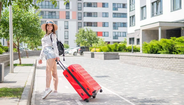 Mooie jonge blanke vrouw in een zomerhoed poserend met een rode koffer buiten. Gelukkig lachend meisje gaat op vakantie met een grote bagage tas. Charmante dame in korte broek op een hete zonnige dag. — Stockfoto