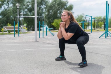 Şişman kız spor sahasında spor yapıyor. Güzel, genç, gülümseyen, kilolu bir kadın dışarıda çömeliyor. Kötü yeme alışkanlıkları. Kalça kasları üzerinde egzersiz yapar..