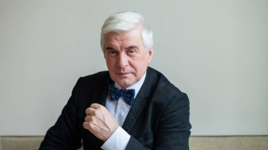 Takım elbiseli ve papyonlu, gri saçlı, gülümseyen bir adamın portresi. Zarif giyimli, temiz tıraşlı bir son sınıf öğrencisi kameraya özgüvenle bakıyor..