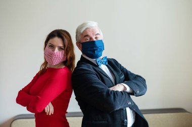 Takım elbiseli olgun gri saçlı bir adam ve kırmızı elbiseli orta yaşlı bir kadın sırt sırta verip tıbbi maskeler takıyorlar. Sosyal mesafeyi koruyan meslektaşların portresi.
