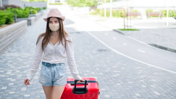 Een mooie jonge blanke vrouw met een medisch masker loopt door de straat met een rode koffer. Een meisje met bagage reist alleen tijdens de hoogte van de coronavirusepidemie. De lege stad. — Stockfoto