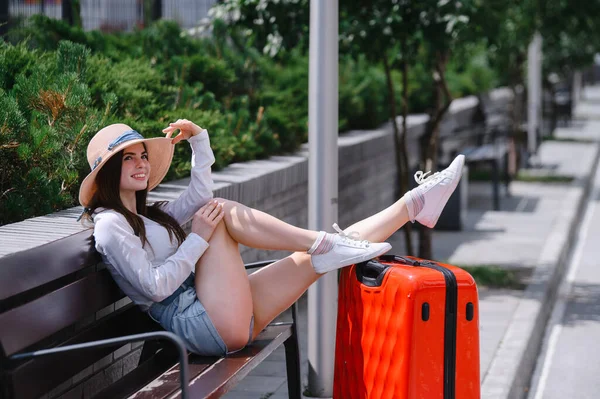 Mooie brunette in korte broek en een hoed zit op een bank gestrekte benen op een grote rode koffer. Blanke vrouw vouwde haar voeten op een tas buiten. Meisje geniet van een reis in het park op een zonnige. — Stockfoto