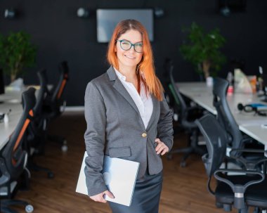 Takım elbiseli ve gözlüklü güzel kızıl saçlı iş kadını modern bir ofiste dizüstü bilgisayar tutuyor. Başarılı bir kadın açık bir uzay ofisinde çalışıyor. Büyük bir şirketin kendine güvenen kadın lideri..