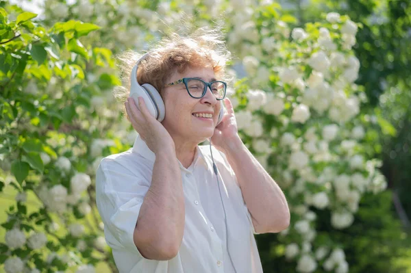 Glad gammal kvinna går i parken av blommande äppelträd och lyssnar på musik på hörlurar. En kvinnlig pensionär ler och njuter av en varm sommardag i trädgården. Avancerad äldre generation. — Stockfoto