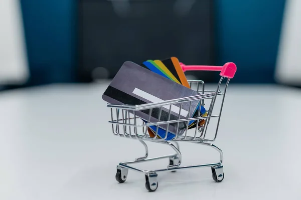 Um mini carrinho de compras cheio de cartões de crédito fica em uma mesa branca. Não há pessoas. Close-up de um carrinho com vários cartões de débito bancários diferentes . — Fotografia de Stock