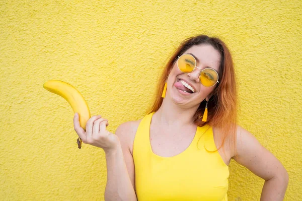 Красивая рыжеволосая женщина в жёлтом платье и солнечных очках показывает язык и держит банан. Портрет игривой девушки, фантазирующей с спелыми фруктами. Леди с прекрасной ослепительной улыбкой . — стоковое фото
