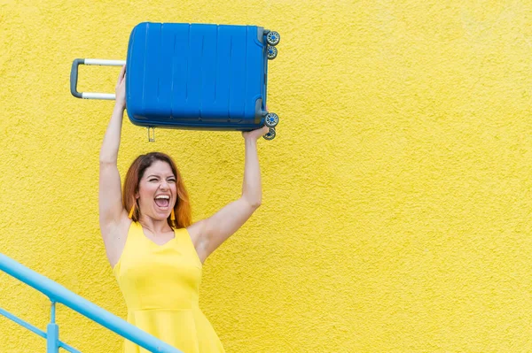Happy woman in a yellow dress holds a blue suitcase over her head against the background of a yellow wall. Caucasian girl smiles with teeth and happily waits for a trip.