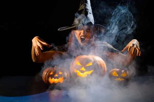 Złowroga wiedźma w kapeluszu przywołuje latarnię. Tradycyjne postacie halloween. Mistyczna mgła skrada się nad dyniami z wyrzeźbionymi okropnymi twarzami. Kobieta w stroju karnawału. — Zdjęcie stockowe