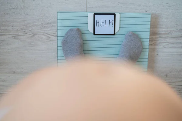 Faceless fat woman measures weight on a bathroom scale. Top view of a large naked female belly and feet in gray socks on the scales with the word help. The inscription on the screen help. Obesity.