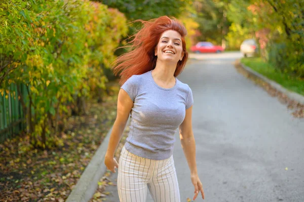 아름다운 빨간 머리의 여자가 거리를 걷고 춤을 추고 있습니다. 회색 티셔츠에 줄무늬 레깅스를 입은 행복 한 소녀는 가을 파크에서 산책을 즐기고 있다. 움직이는 사진. — 스톡 사진