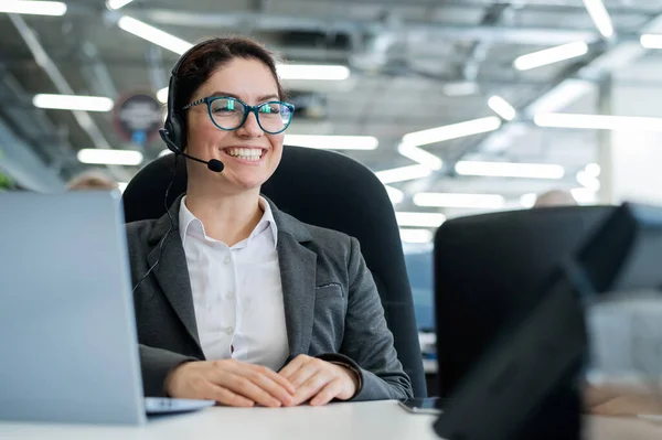 A happy female support operator is sitting at a desk and answering calls. Beautiful smiling woman talking to customers on a headset. Office employee in headphones.