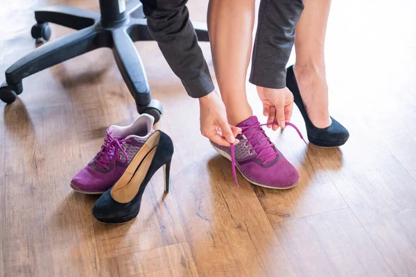 Крупный план ног офисной работницы с болью в ноге. Женщина меняет высокие черные замшевые каблуки на более удобные туфли. Девушка снимает обувь и надевает фиолетовые кроссовки.. — стоковое фото